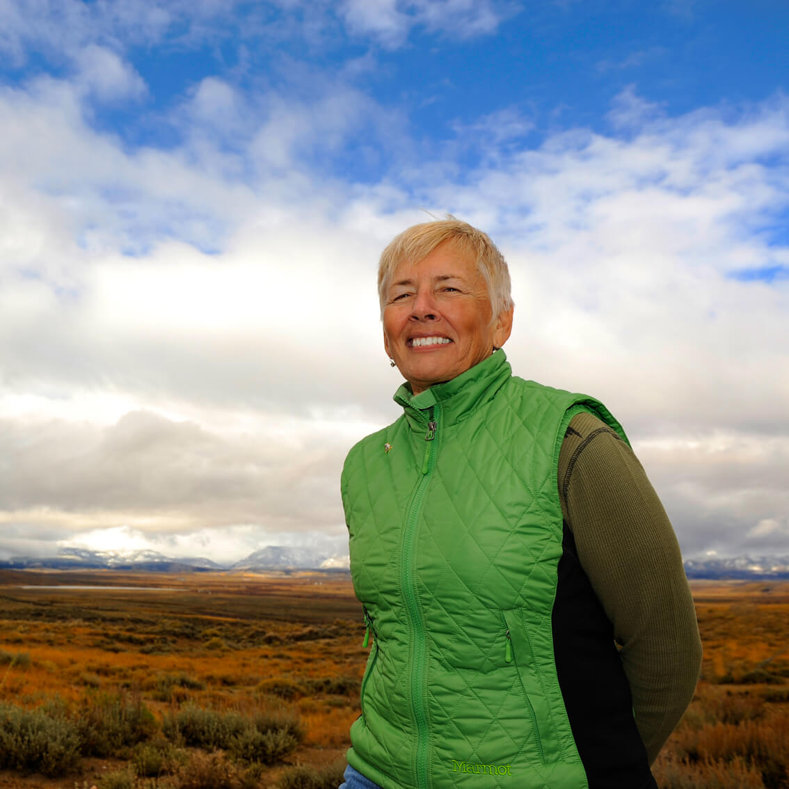 Person in a green vest stands outdoors with a vast landscape in the background, partially cloudy sky overhead.