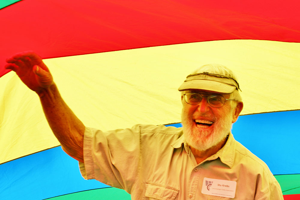 A photo portrait of Stu Krebs, laughing and holding his hand up in front of a rainbow-colored banner