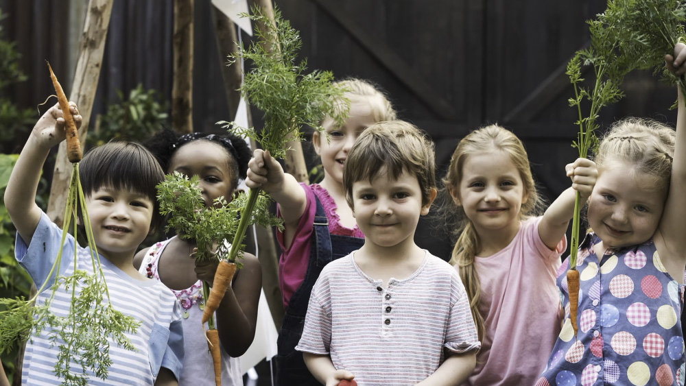 Children smile at the camera and hold up vegetables