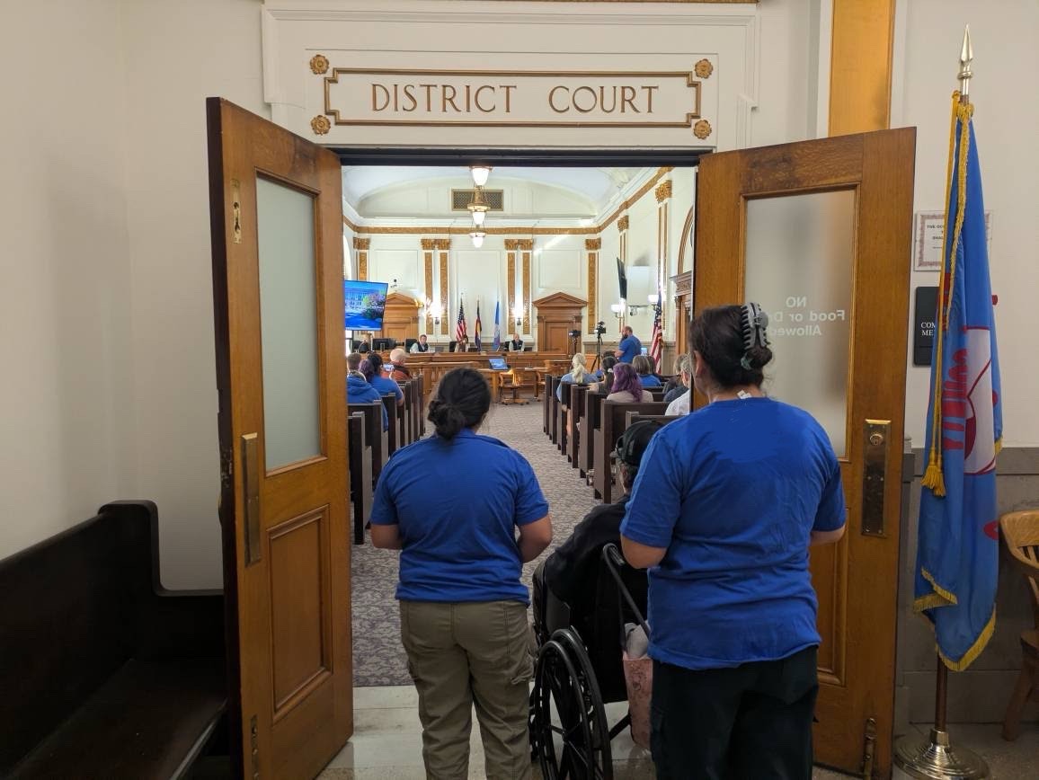 People in Western Colorado Alliance t-shirts stand outside the courthouse where a WCA member is addressing County Commissioners