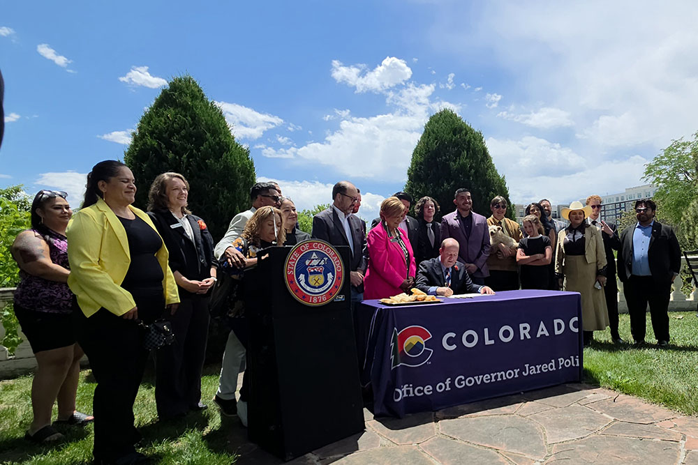 Governor Jared Polis signs the Tamale Act at an outdoors table surrounded by the bill's sponsors and other affected Coloradans
